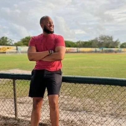 Man standing outdoors near a chain-link fence, looking to the side with arms crossed, on a grassy field under a partly cloudy sky.