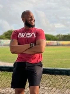 A man wearing a red NASA t-shirt and black shorts stands outdoors with arms crossed, smiling and looking to the side. Background features a grassy field, a fence, and partly cloudy sky.