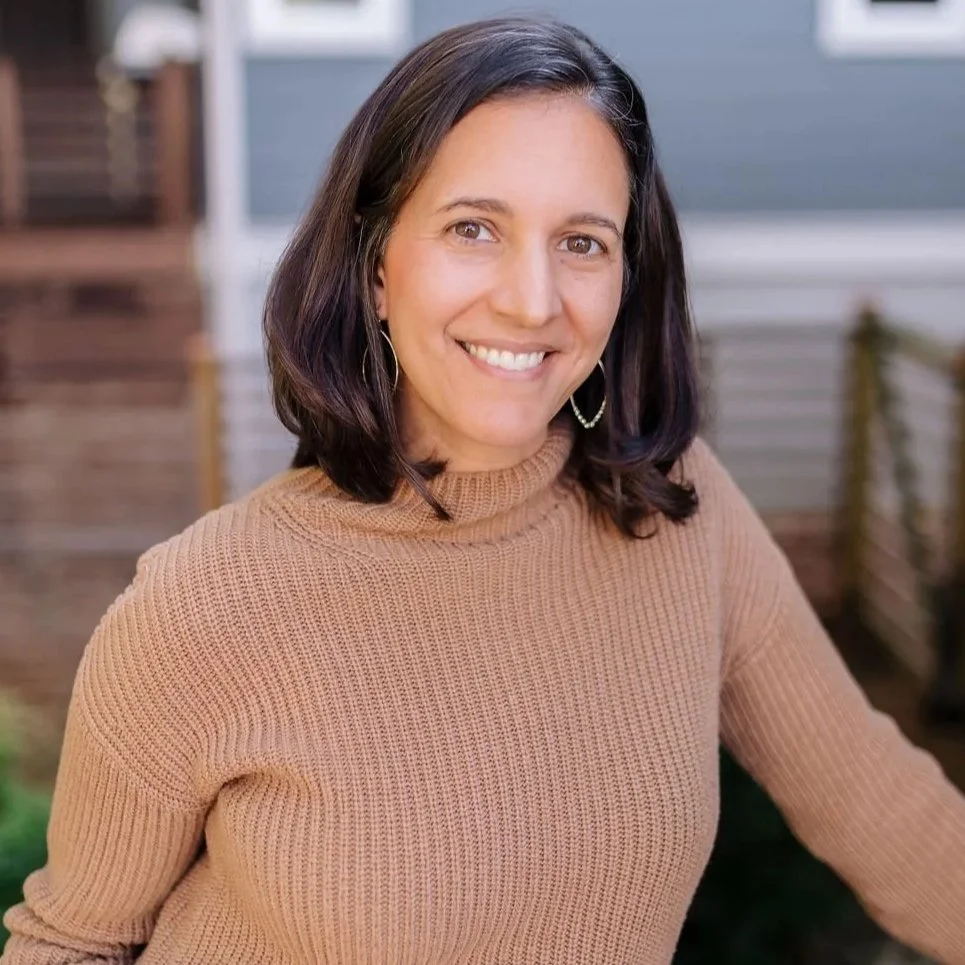 Therapist Keely Clark LCSW, PMH-C with shoulder-length dark hair, wearing a tan knit sweater and hoop earrings, smiling warmly outdoors in front of soft blue backdrop