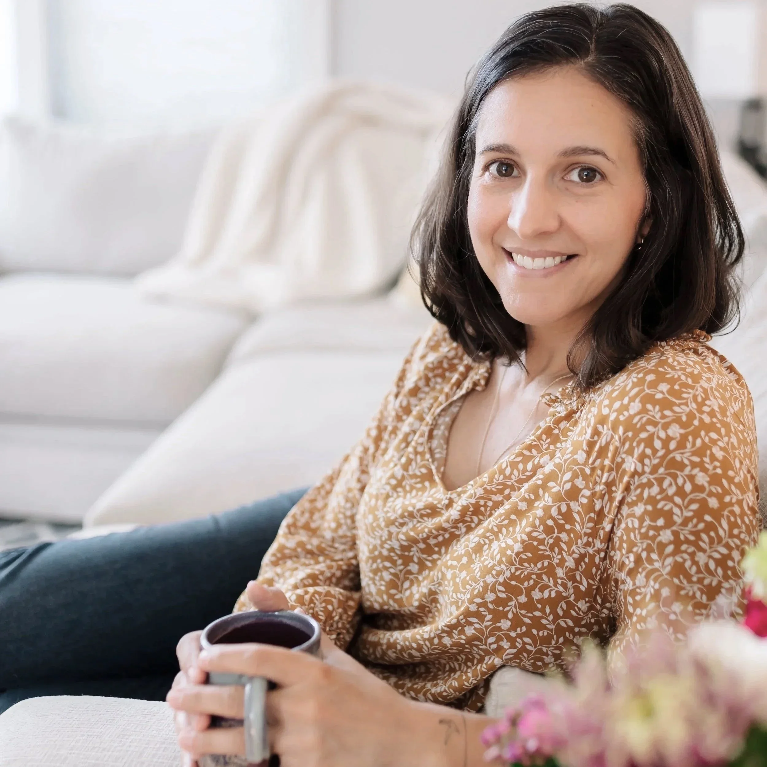 A woman with shoulder-length brown hair smiling and holding a mug, sitting on a beige sofa in a bright living room.