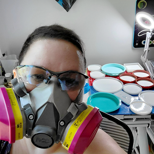 Artist Ava Bock wearing a fume rated safety mask in her studio, standing in front of circular molds filled with polyurethane and bio-resin epoxy as part of her careful and health-conscious art-making process.