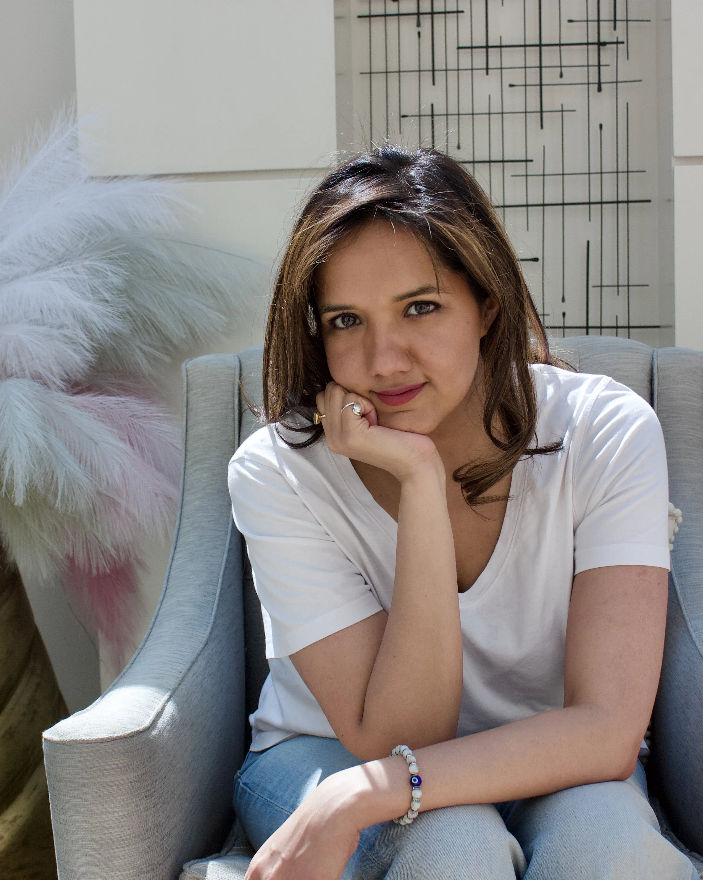 Vidhyah Coaching Founder and Coach Shruti Agrawal - sitting on a light coloured red armchair, resting her chin on her hand, wearing a white t-shirt, blue jeans, and jewellery. Behind her is abstract wall art and a feathered decoration.