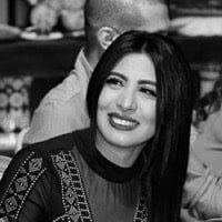 A woman smiling with a man in the background, both outside.