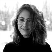 A woman with shoulder-length curly hair and glasses smiling, wearing a dark top.