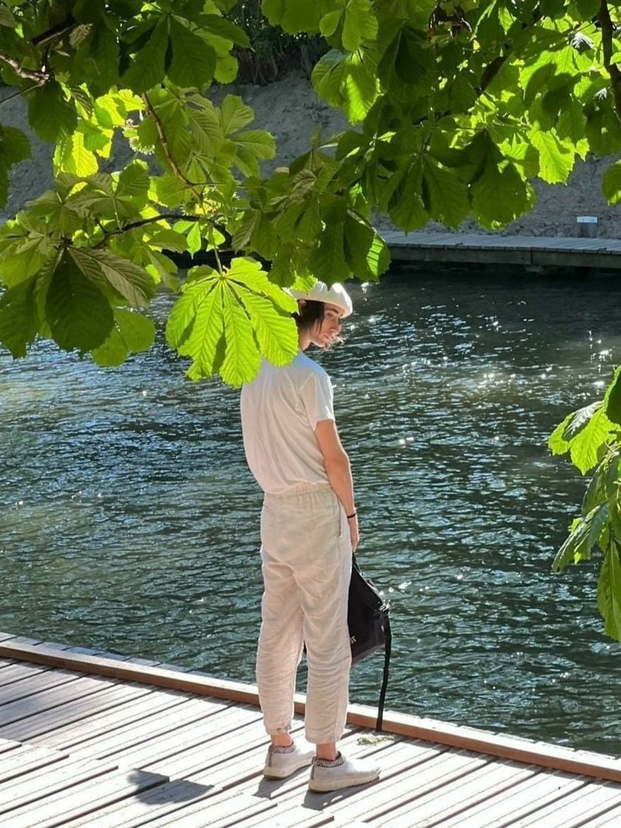 A woman standing on a wooden dock by a body of water, wearing a white hat, white t-shirt, beige pants, and holding a black bag, partially obscured by large green leaves in the foreground.