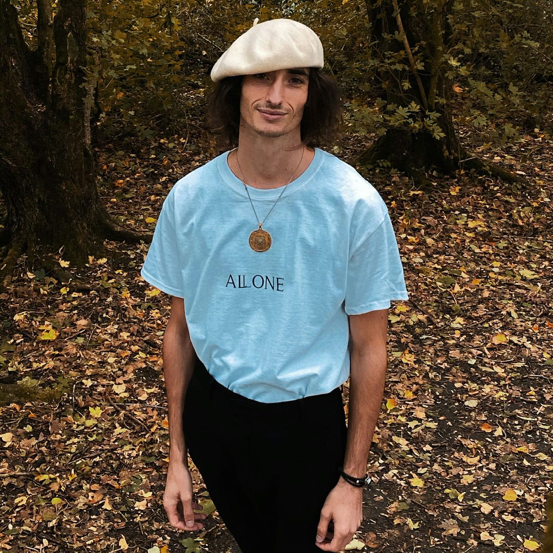 A man with long dark hair wearing a beige beret, a light blue T-shirt with the word 'ALLONE' printed on it, black pants, and a necklace with a circular pendant, standing in a forest with fallen leaves.