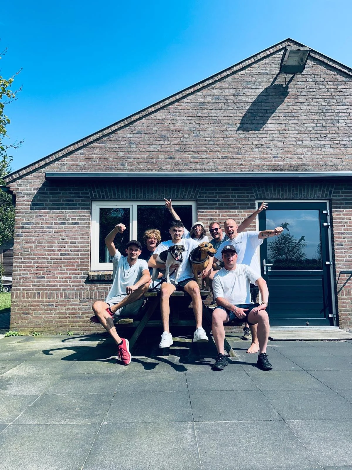 Group of seven people with two dogs sitting and standing on a wooden bench in front of a brick house with large windows and a door, celebrating outdoors on a sunny day.