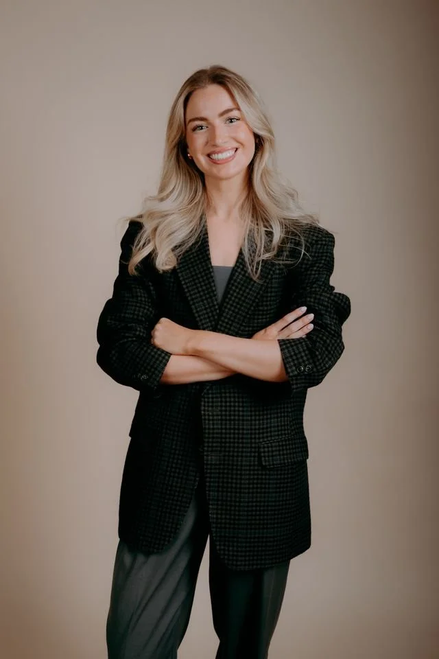 A smiling young woman with blonde hair wearing a black shirt and safety glasses around her neck standing against a plain light-colored wall.