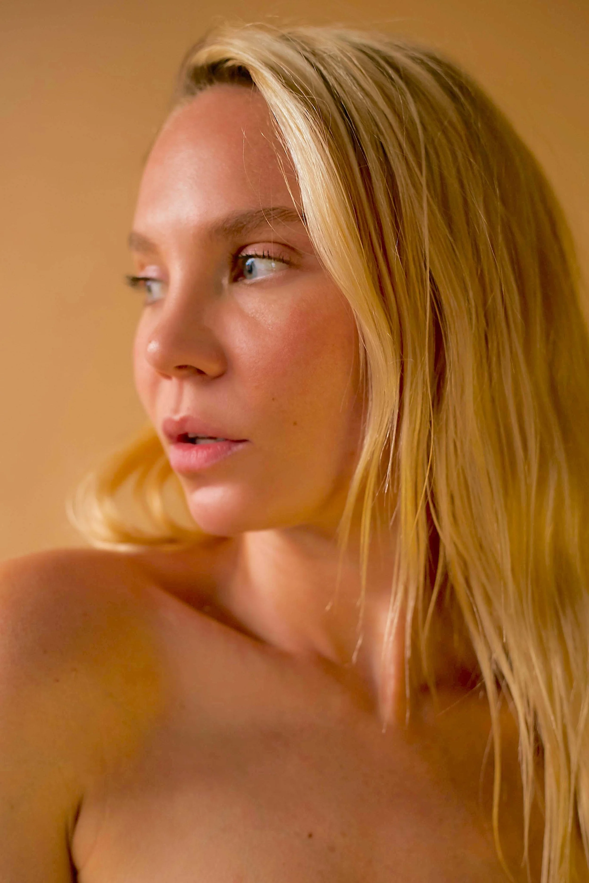 A close-up of a blonde woman with blue eyes looking to her left, with a warm, neutral background.