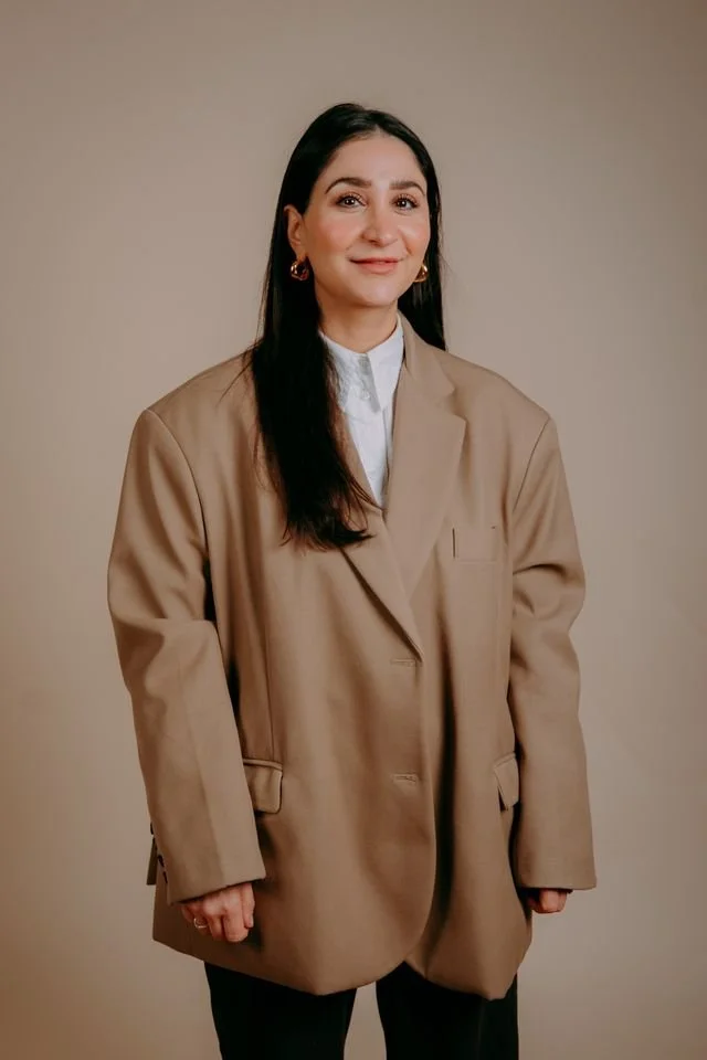 A woman with long dark hair, wearing a white oversized shirt, seated on a wooden stool in front of a pink backdrop. She is crossing her legs, wearing white sneakers with black stars, and looking to her left with a slight smile.
