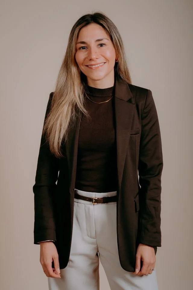 A woman with long blonde hair smiling, wearing a black blazer, black top, white pants, and a gold necklace, standing against a plain beige background.
