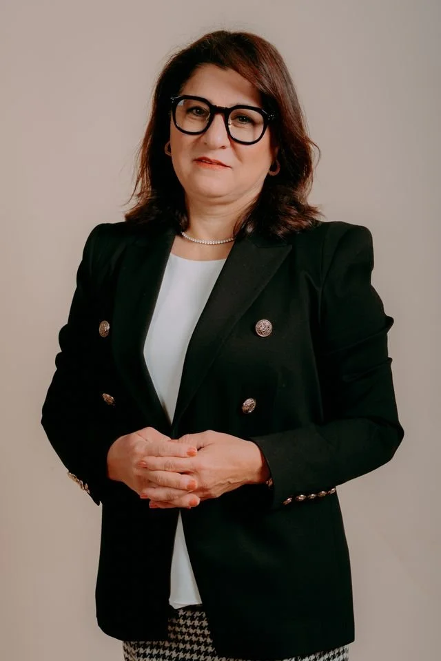 A woman with shoulder-length dark hair wearing black glasses, a black blazer with metallic buttons, a white top, and a pearl necklace, standing against a plain background.