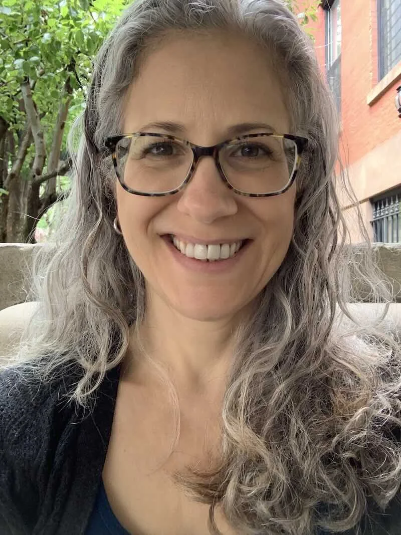 Carol Covelli, LCSW, smiling and wearing tortoiseshell glasses, with long gray wavy hair, outdoors in front of green trees and a brick building.