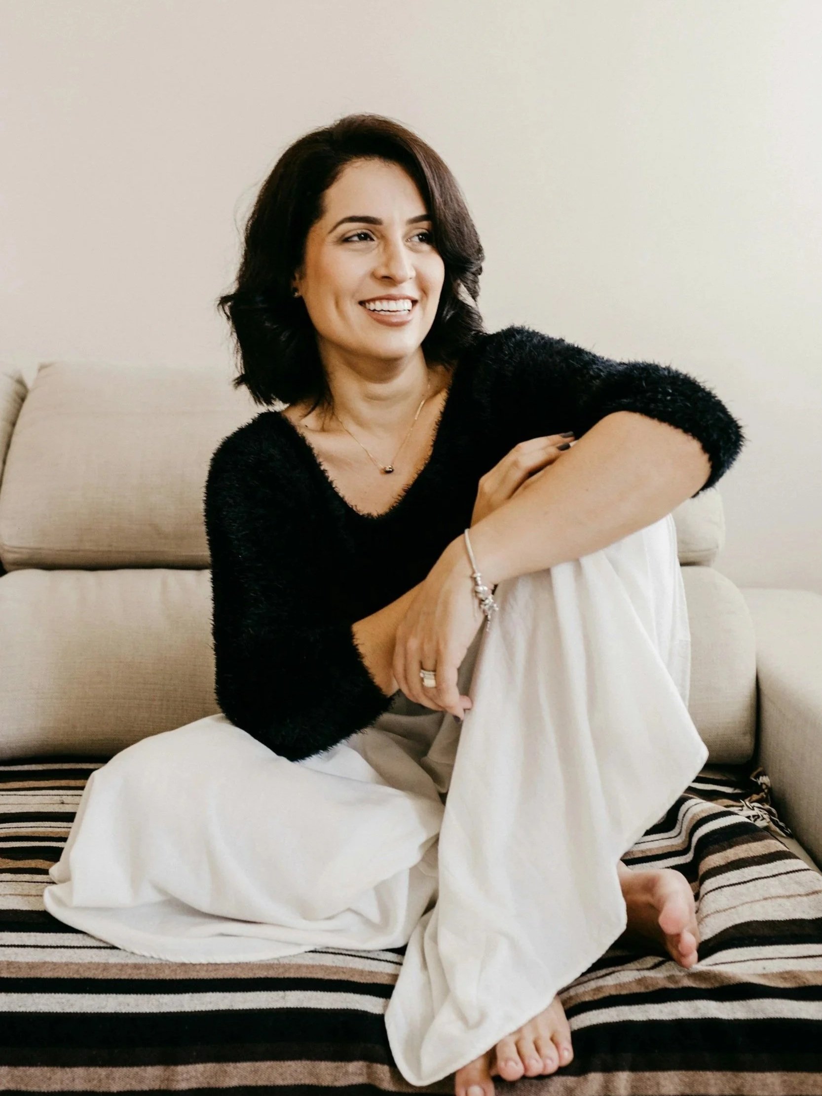 A woman with dark, shoulder-length hair, wearing a black fuzzy sweater and white pants, sitting barefoot on a striped bed or couch, smiling and looking to her left.