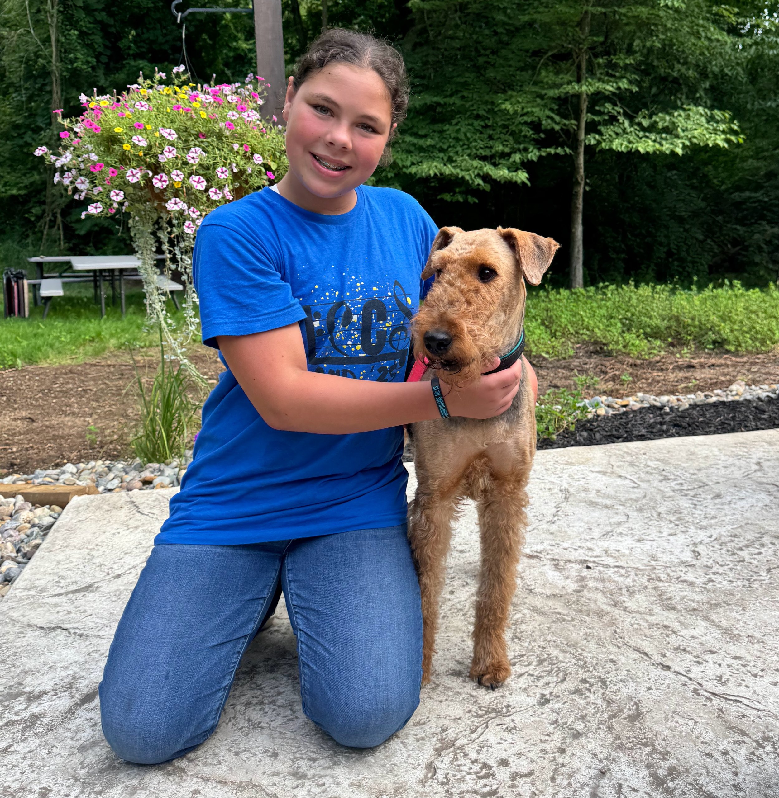 A young girl kneels on the ground outside with her arms around an Airedale Terrier. They are both smiling.