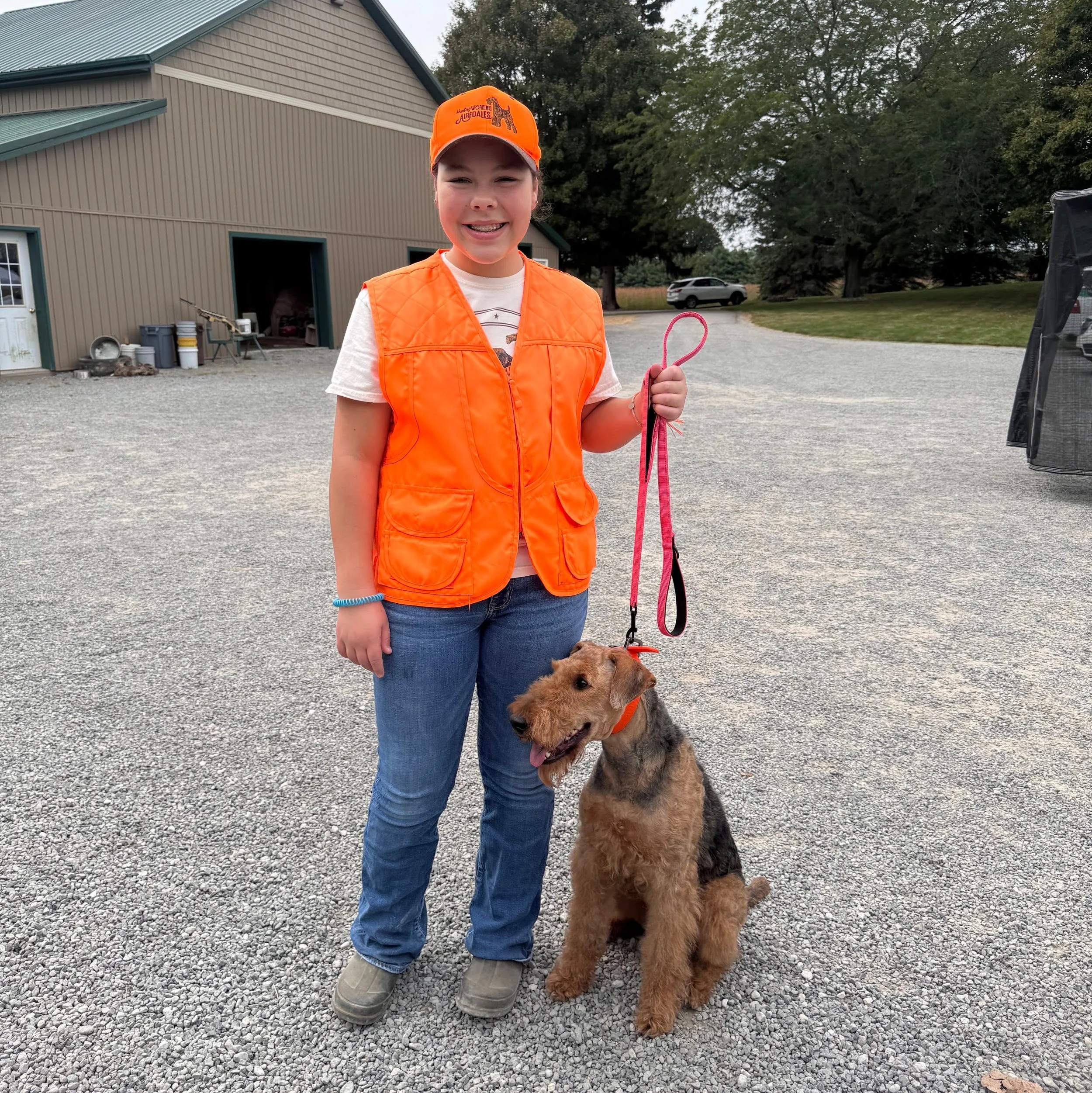 Young girl in an orange hunting vest and hat holding a pink leash attached to a brown and black Airedale dog sitting on a gravel driveway with a hunt club in the background.