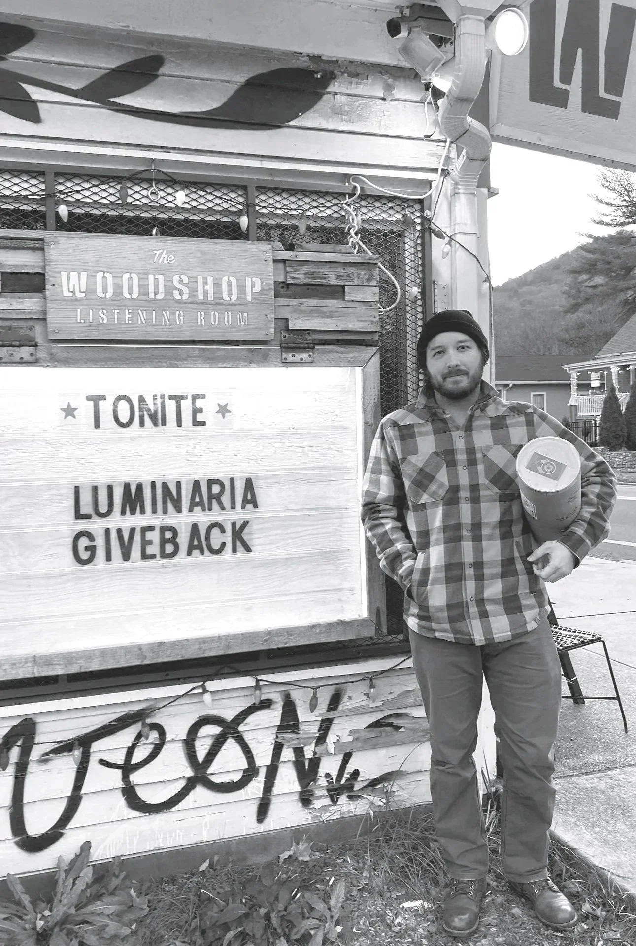 A grayscale photograph of photographer Kenny Thatcher outside the Woodshop Listening Room holding a large print tube, next to the marquis which reads "Tonight - Luminaria Giveback", and has a bit of grafiti along the bottom of the building.