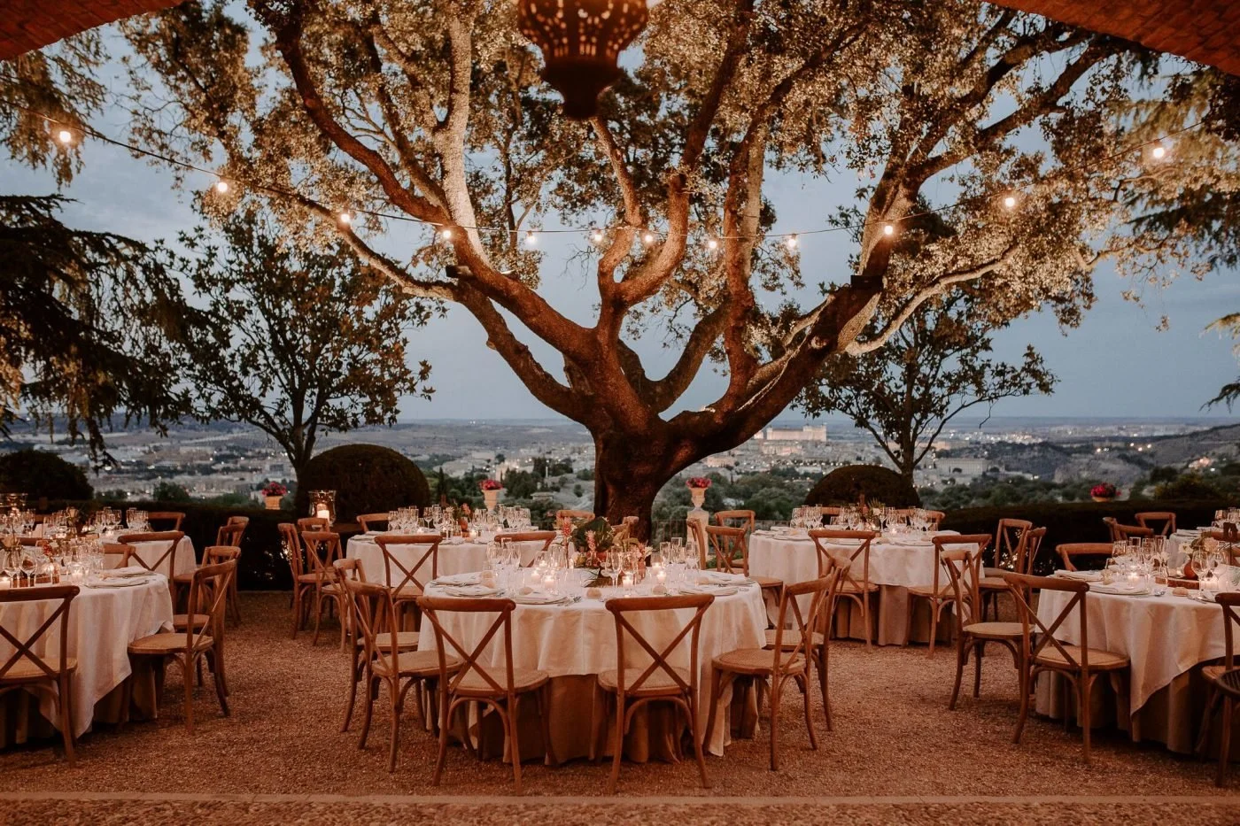 Romantic outdoor wedding reception with candlelit tables beneath a large tree at dusk.