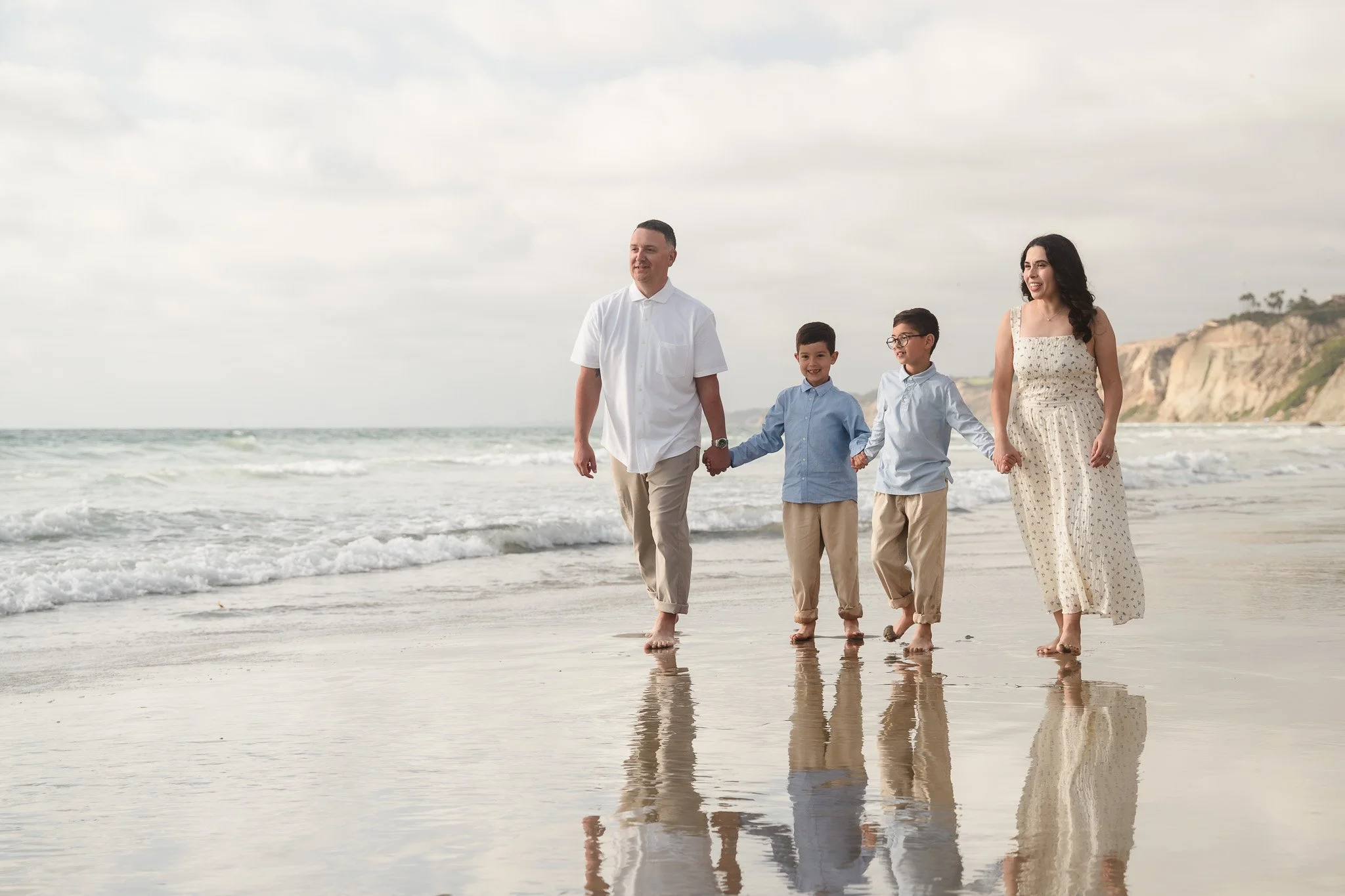 A family of four walking hand in hand along the shoreline at the beach, with a cloudy sky and cliffs in the background.