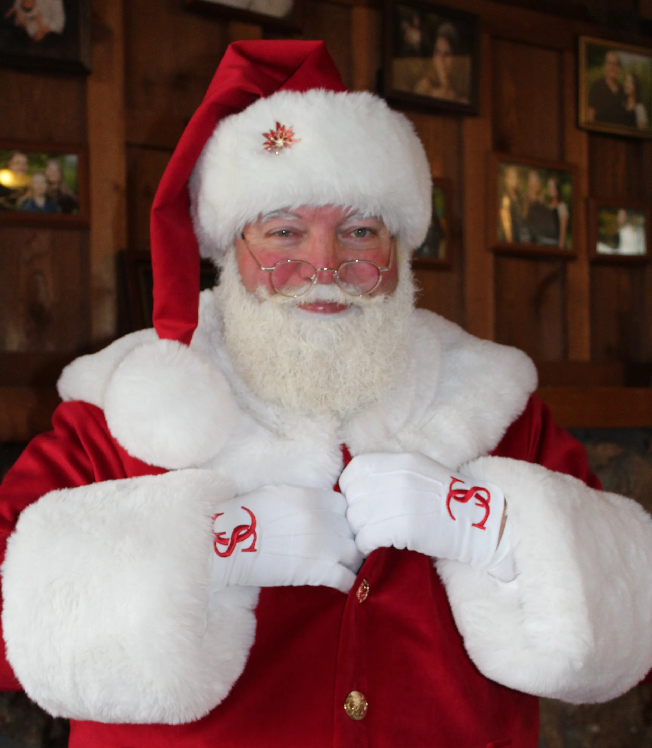 Santa Claus in a traditional red and white suit with glasses, white gloves, and a white beard, standing indoors with framed pictures in the background.