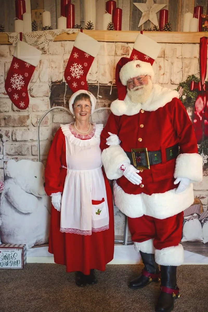 A cheerful couple dressed as Santa Claus and Mrs. Claus stand in front of a cozy fireplace with festive stockings and candles. They are smiling at the camera in a holiday-themed setting.