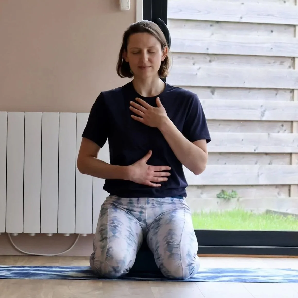 Une femme faisant une séance de méditation par la respiration en position à genoux sur support, une main sur la poitrine et l'autre sur l'estomac.