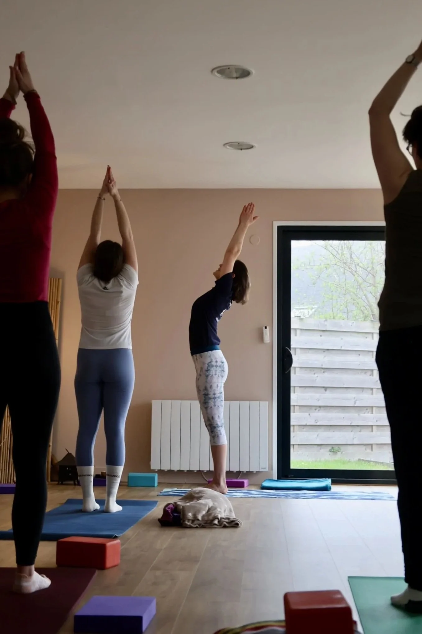 Groupe de personnes pratiquant le yoga dans une pièce lumineuse avec une grande fenêtre arrière, tapis de yoga, blocs.