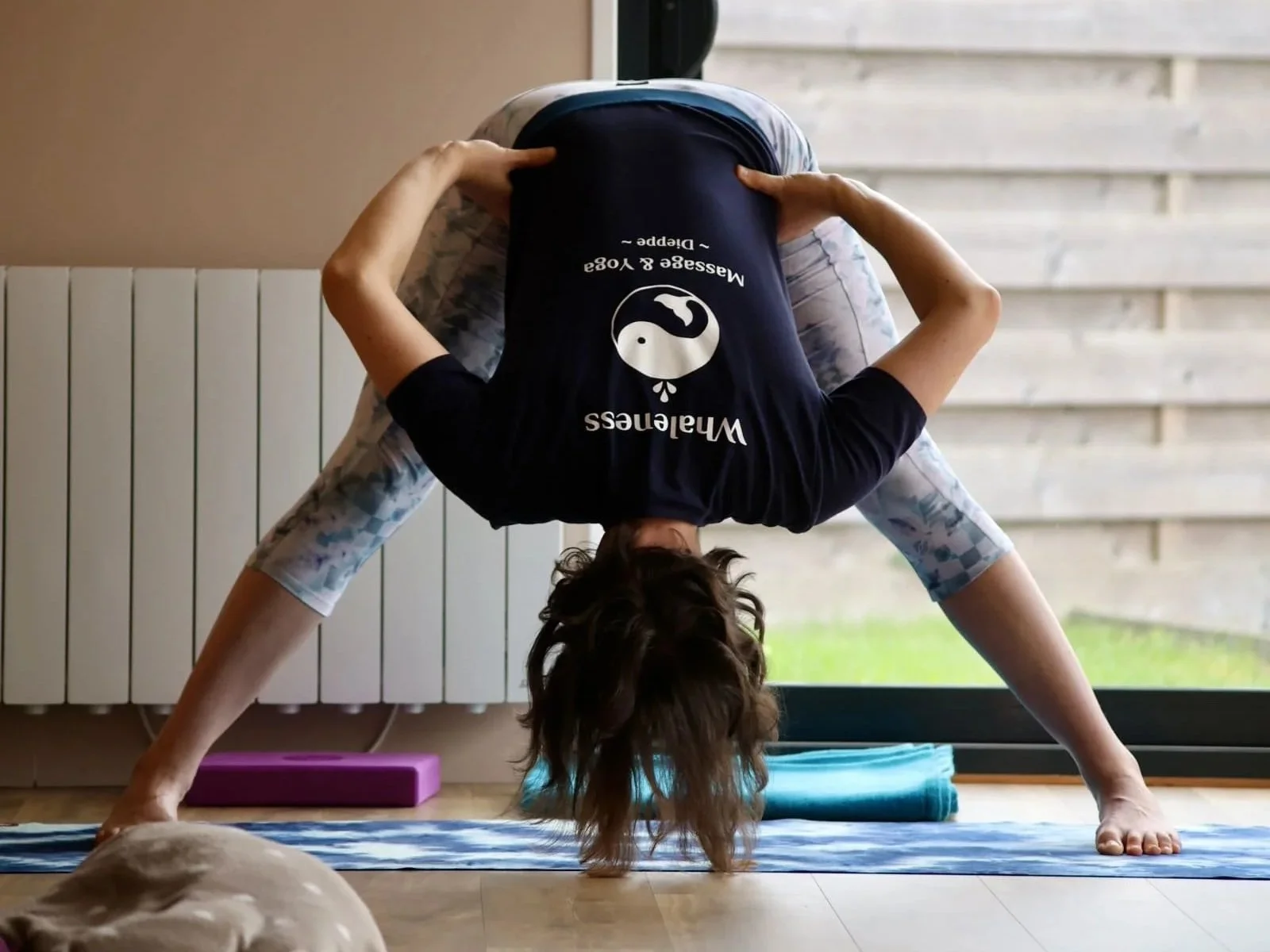 Eugénie, professeure de yoga, pratique dans la salle lumineuse des cours collectifs. Posture de prasarita padottanasana, avec les mains à la taille.