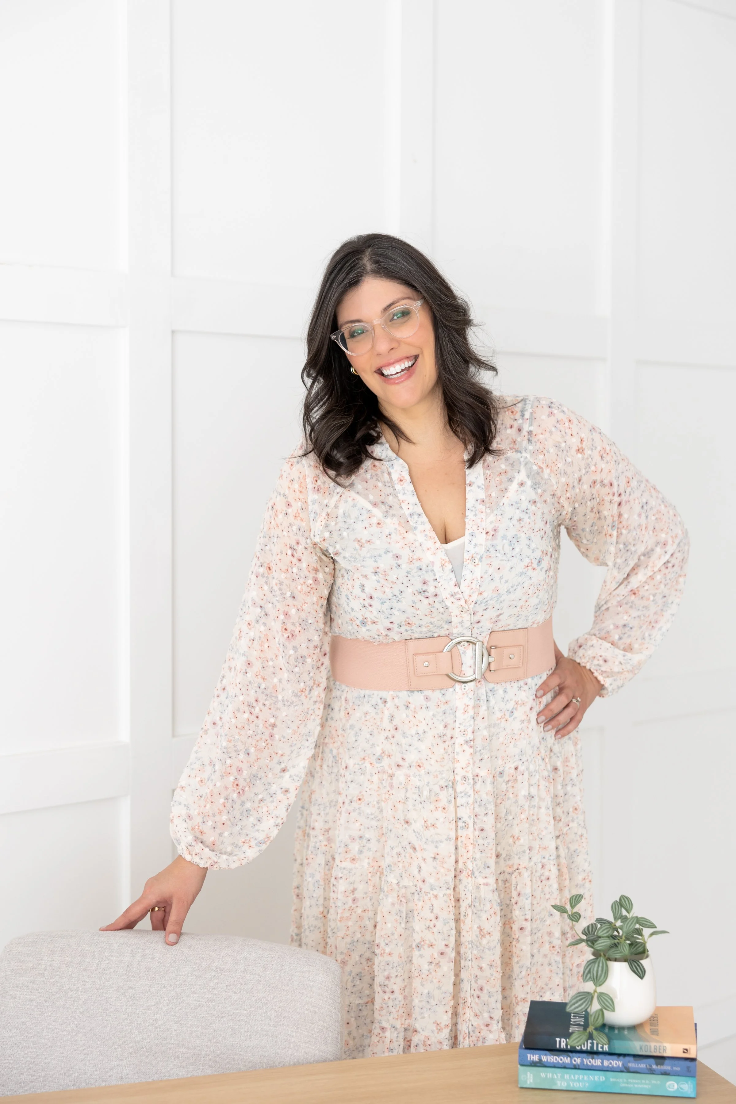 Rebeca Gilbert  smiling, wearing a floral dress with a wide beige belt, standing in a white room near a table with potted plant and stacked books