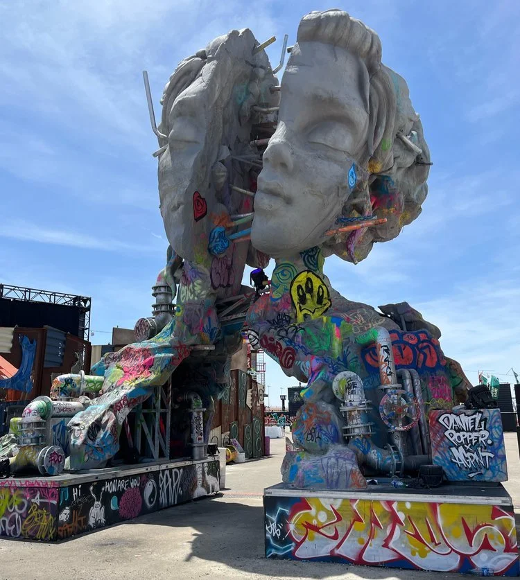 A large outdoor sculpture of a woman's face with multiple heads and graffiti, resembling a mix of a classical statue and street art, set against a blue sky.