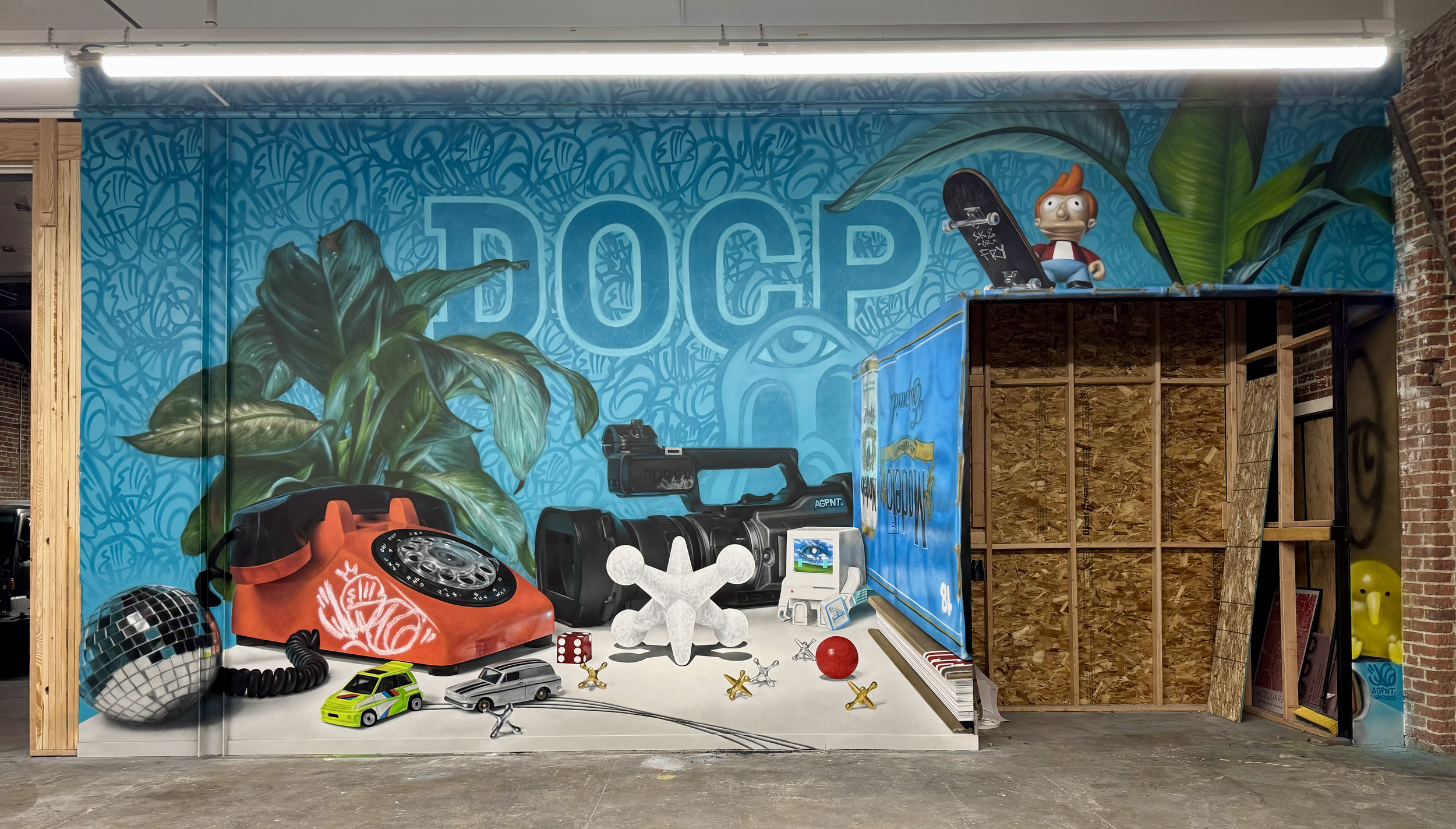 Murals with the word 'DOCP' and various objects including a red rotary phone, a disco ball, toy cars, a drone, a robot, and a cartoon character on a skateboard, with leafy greenery in the background. Still life.