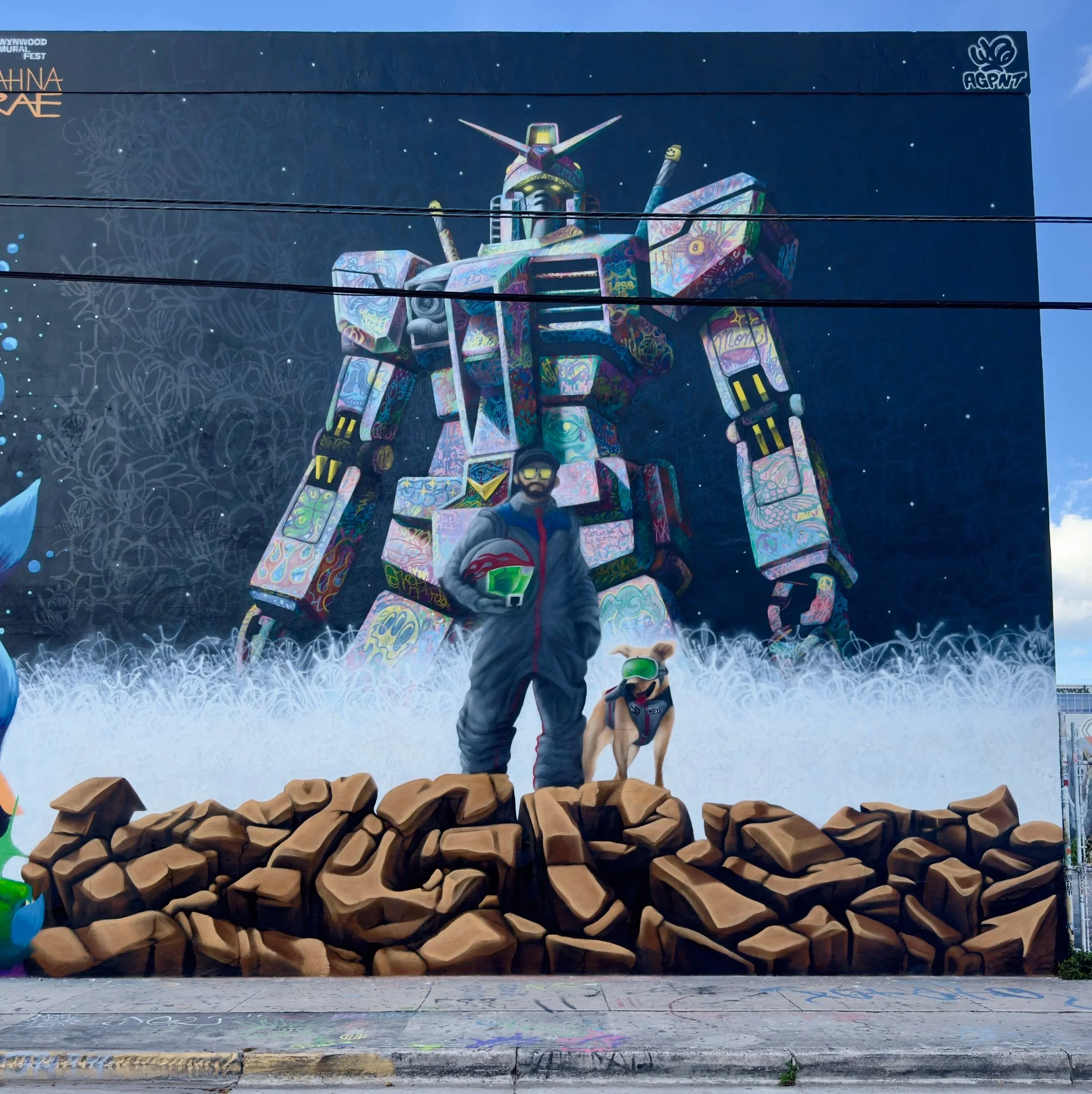 Colorful mural of a giant gundam robot with a human and dog standing on rocks in front of it.