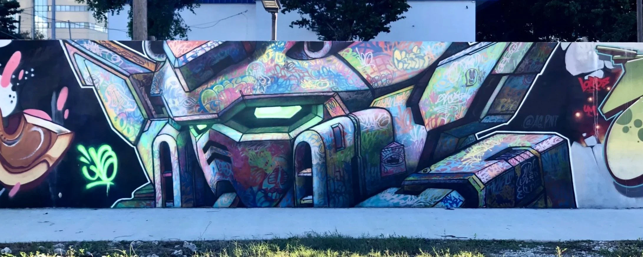 A colorful graffiti mural depicting a robotic or futuristic face with glowing green eyes on a wall outdoors.