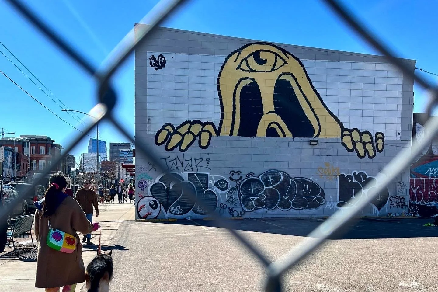 City street view through a chain-link fence sunset mural painting of a large face with an eye on its forehead and hands on the wall, surrounded by graffiti.