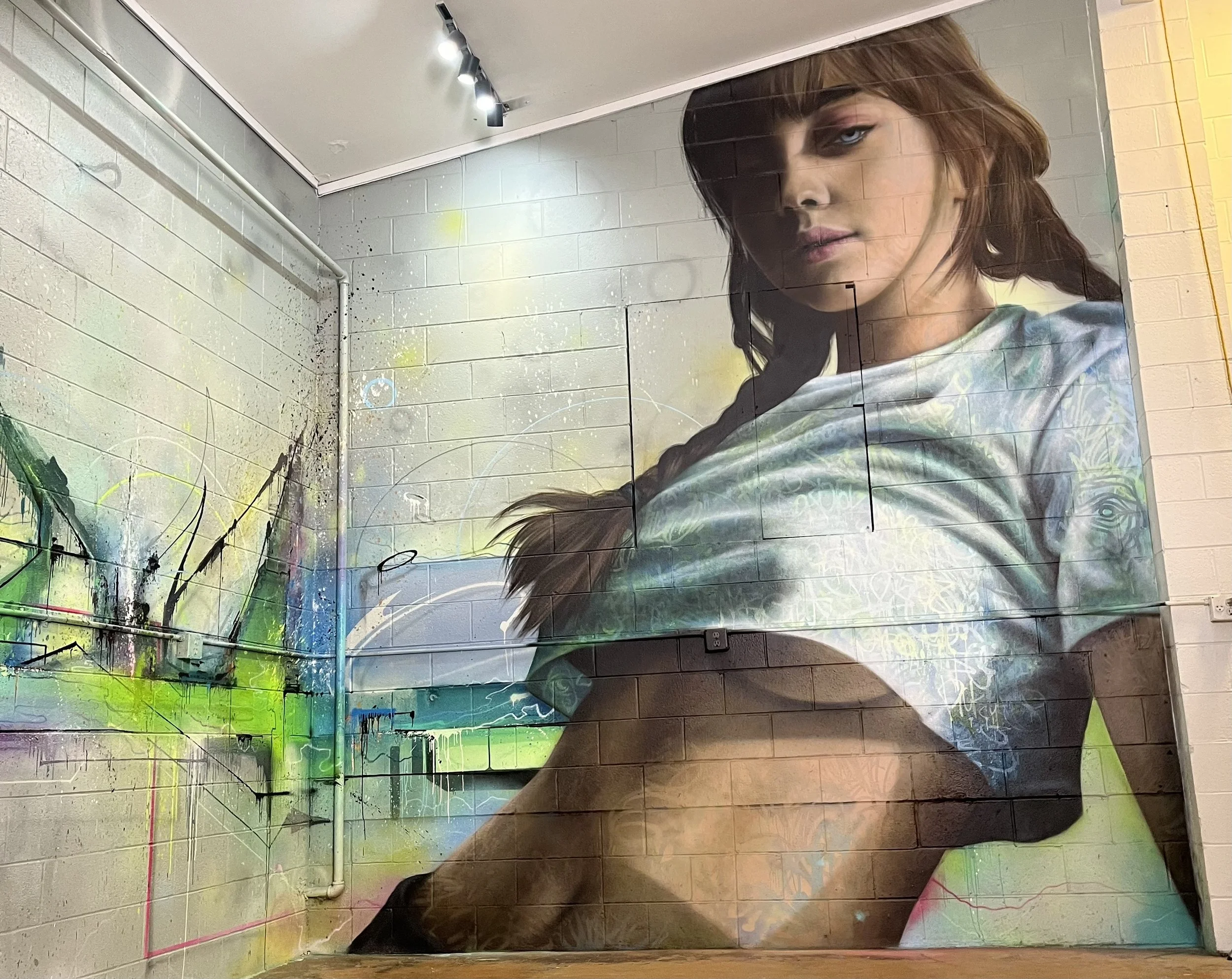 A large mural of a woman with long brown hair, wearing a white shirt and black shorts, sitting on the ground with her knees bent, with abstract and colorful graffiti art on the wall behind her.