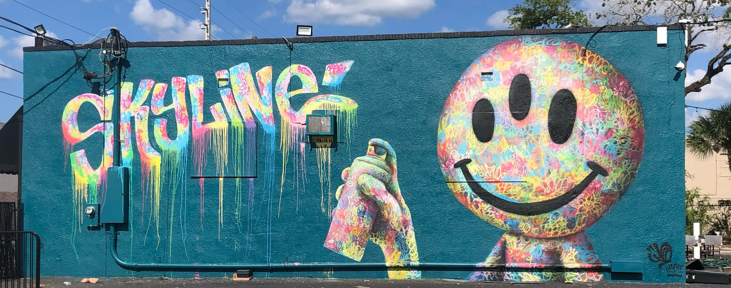 A colorful mural on a blue wall features the word 'SHALOM' in vibrant, multicolored graffiti style, a large smiling face with colorful patterns, and a hand holding a spray paint can with similar colorful designs.