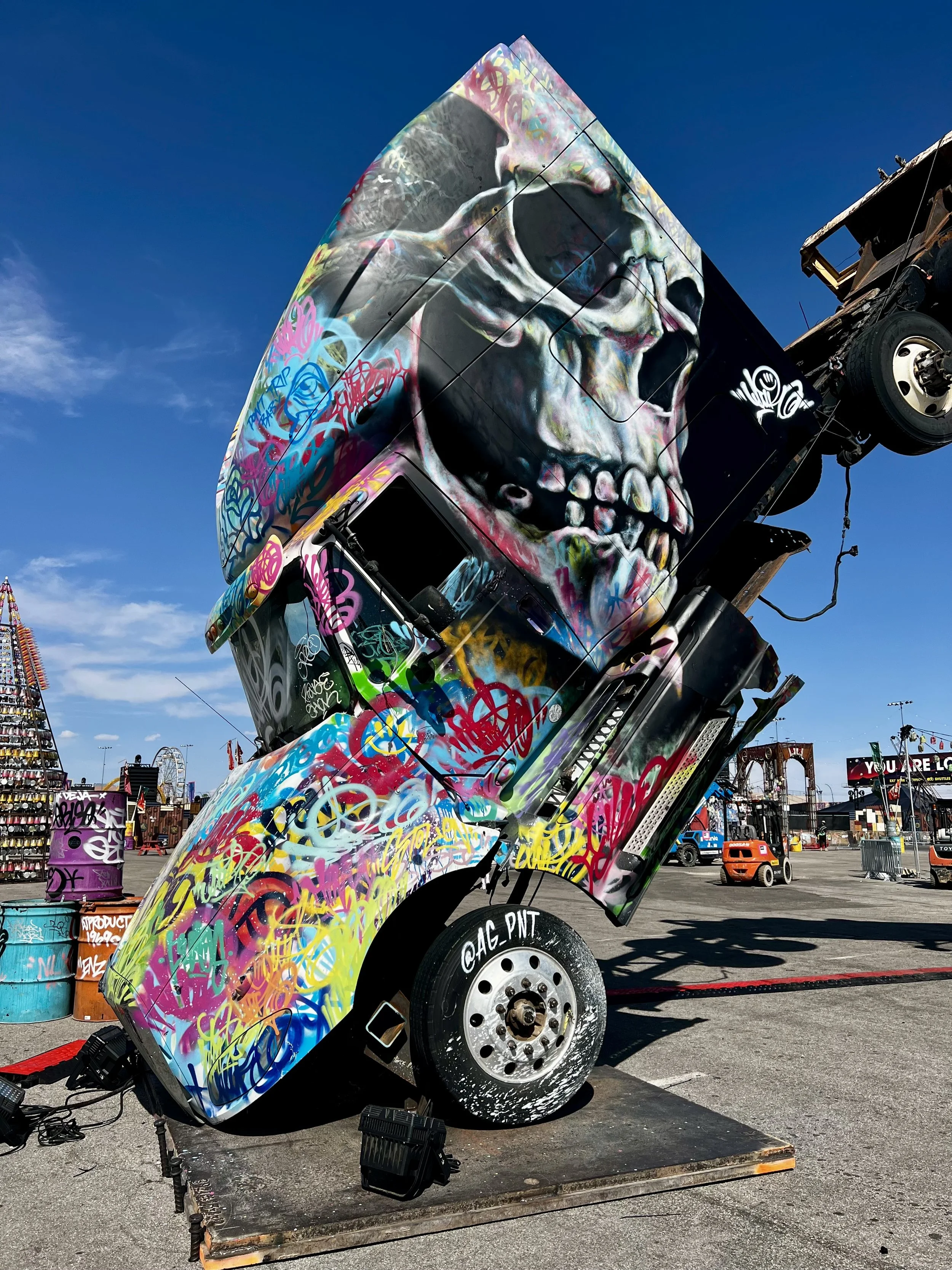 A truck with colorful graffiti art and a large skull painted on the side, tilted on a platform at an outdoor carnival or fairground.