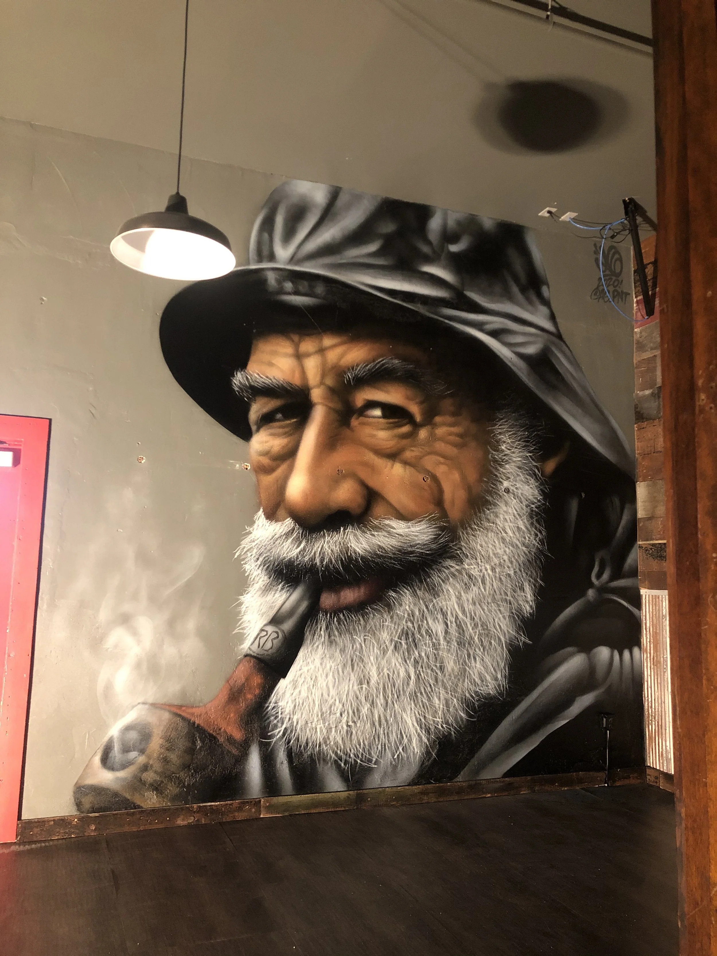 Murale van volwassen man met witte baard en mustache, groot hoofd, rolt sigaret met rook, draagt hoed.