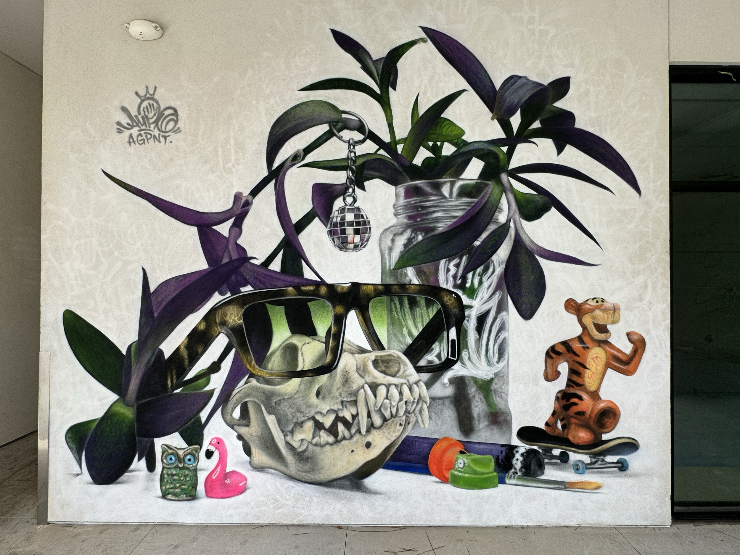 Street art mural with a potted plant, human skull with glasses, a small toy owl, a pink flamingo float, skateboard, skateboard wheels, paintbrush, and a small tiger figure.