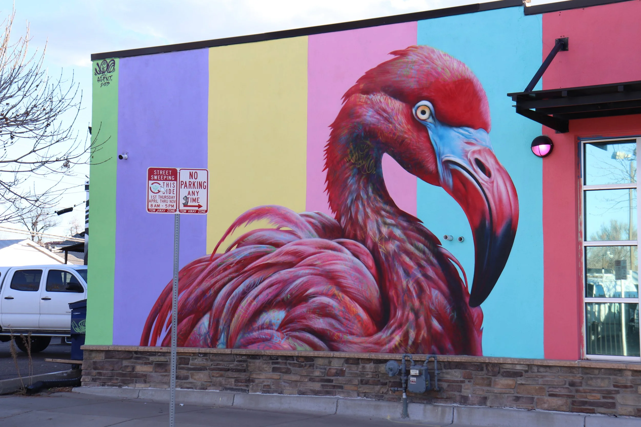 Colorful mural of a flamingo on the side of a building with a pastel rainbow background and street signs in the foreground.