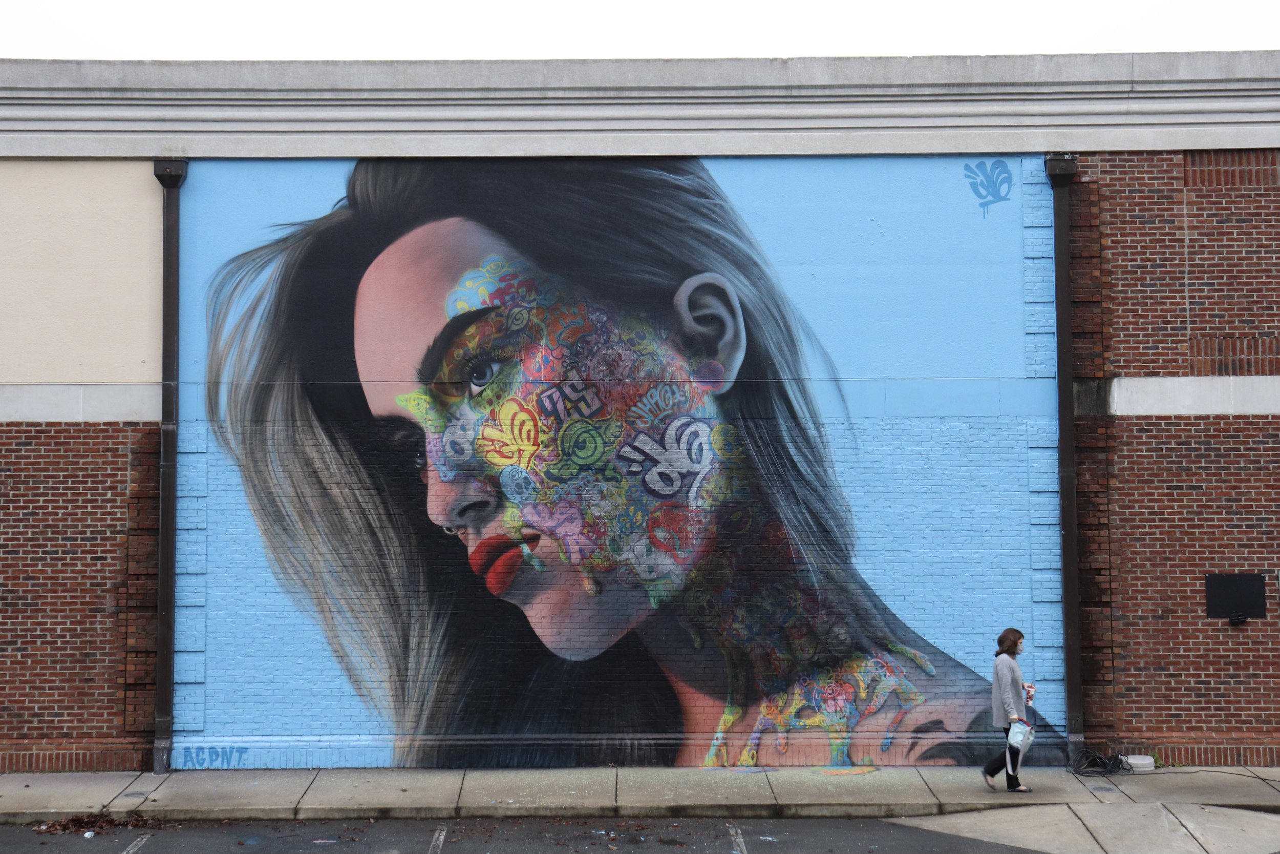 Large mural of a woman's face with colorful graffiti-style artwork on her cheek and neck, painted on a brick building wall, with a person walking by on the sidewalk in front.