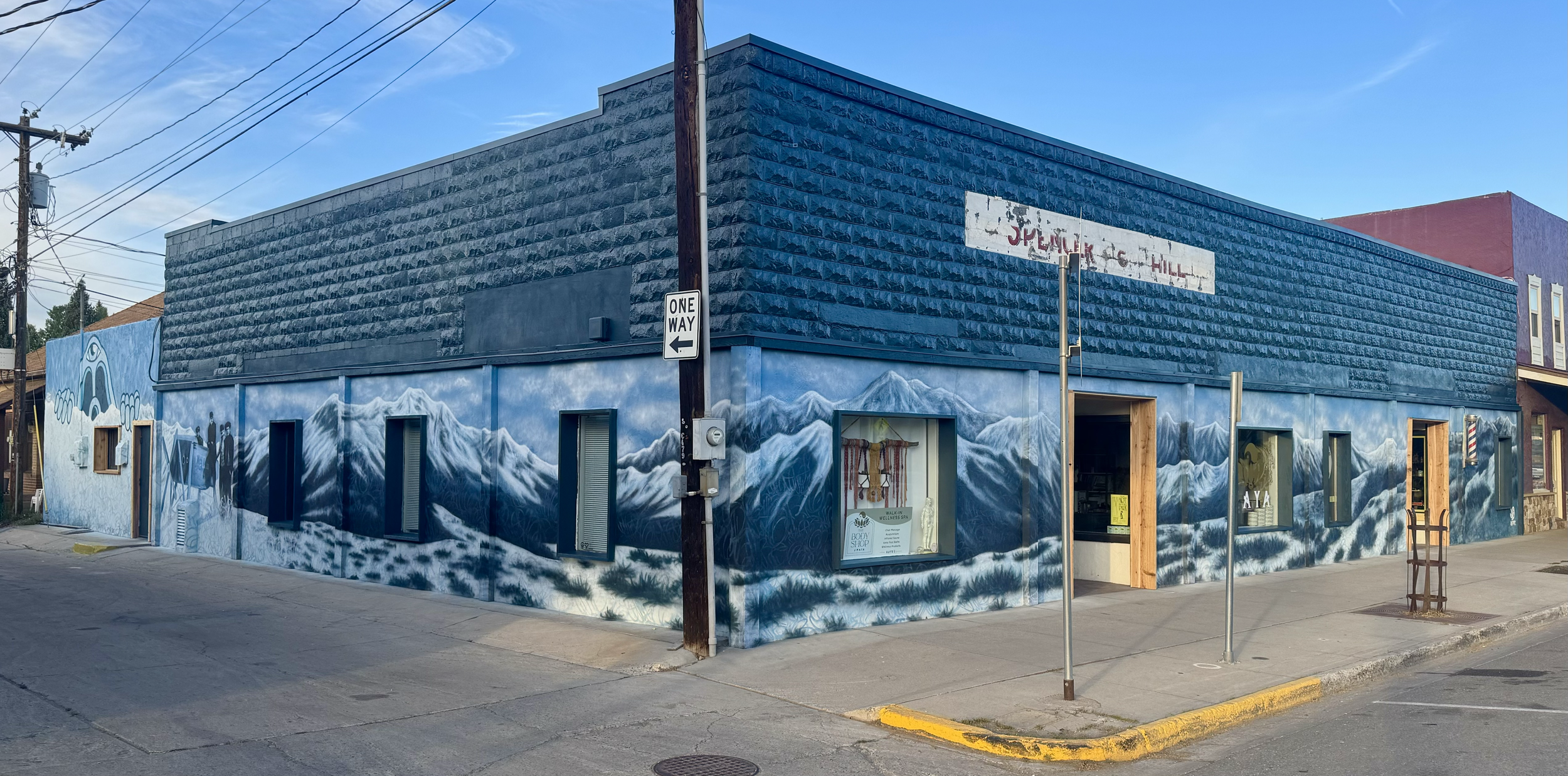 Street corner building with a mural of snow-capped mountains and a sky. The building has windows, some with shutters, and a door. There is a cloudy sky overhead and a pole with a one-way street sign. Part of an adjacent colorful building is visible.