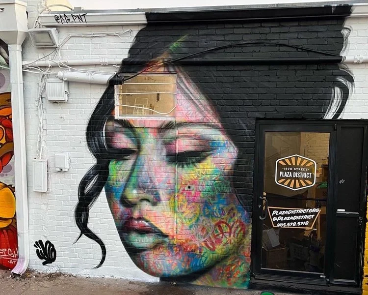 Colorful mural of a woman's face with closed eyes painted on a brick wall outside a building, next to a door with a sign that reads '16th Street Plaza District'.