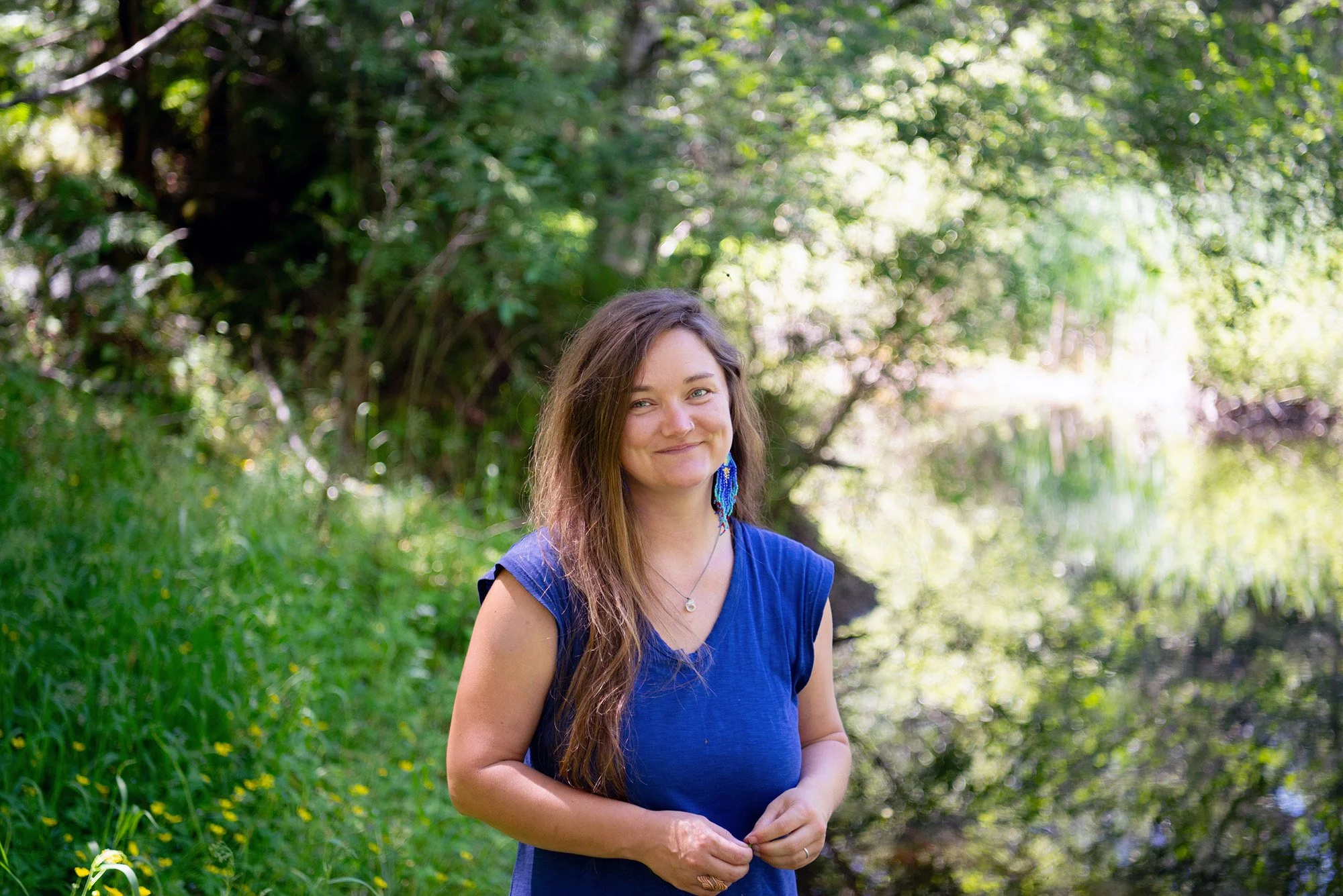 A woman with long brown hair, wearing a blue sleeveless top and blue earrings, stands outdoors near a body of water, surrounded by greenery and trees, smiling softly at the camera.