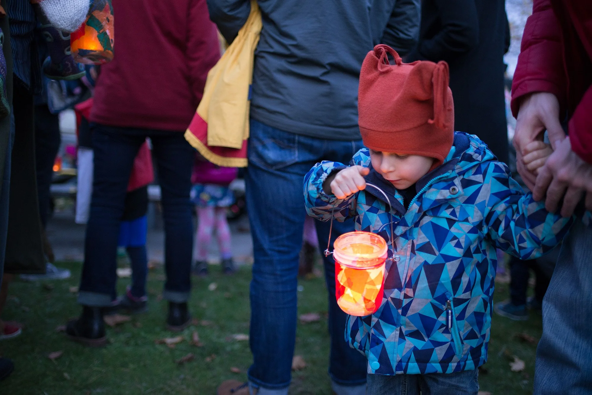 A young child in a blue patterned jacket and red hat looking at a glowing lantern hanging from a string, surrounded by other people in outdoor clothing.