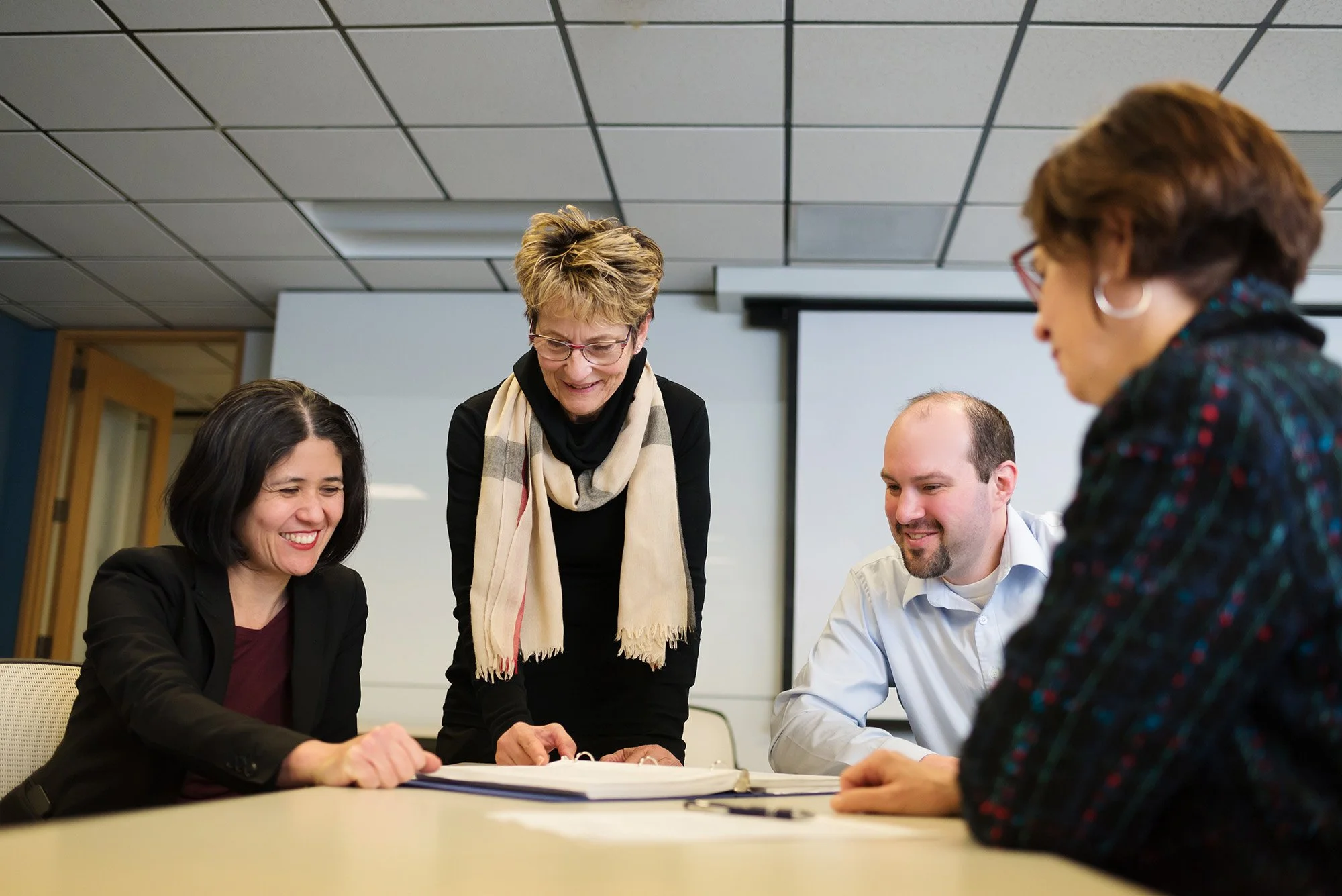 Four coworkers in a meeting room, smiling and looking at a notebook on the table.