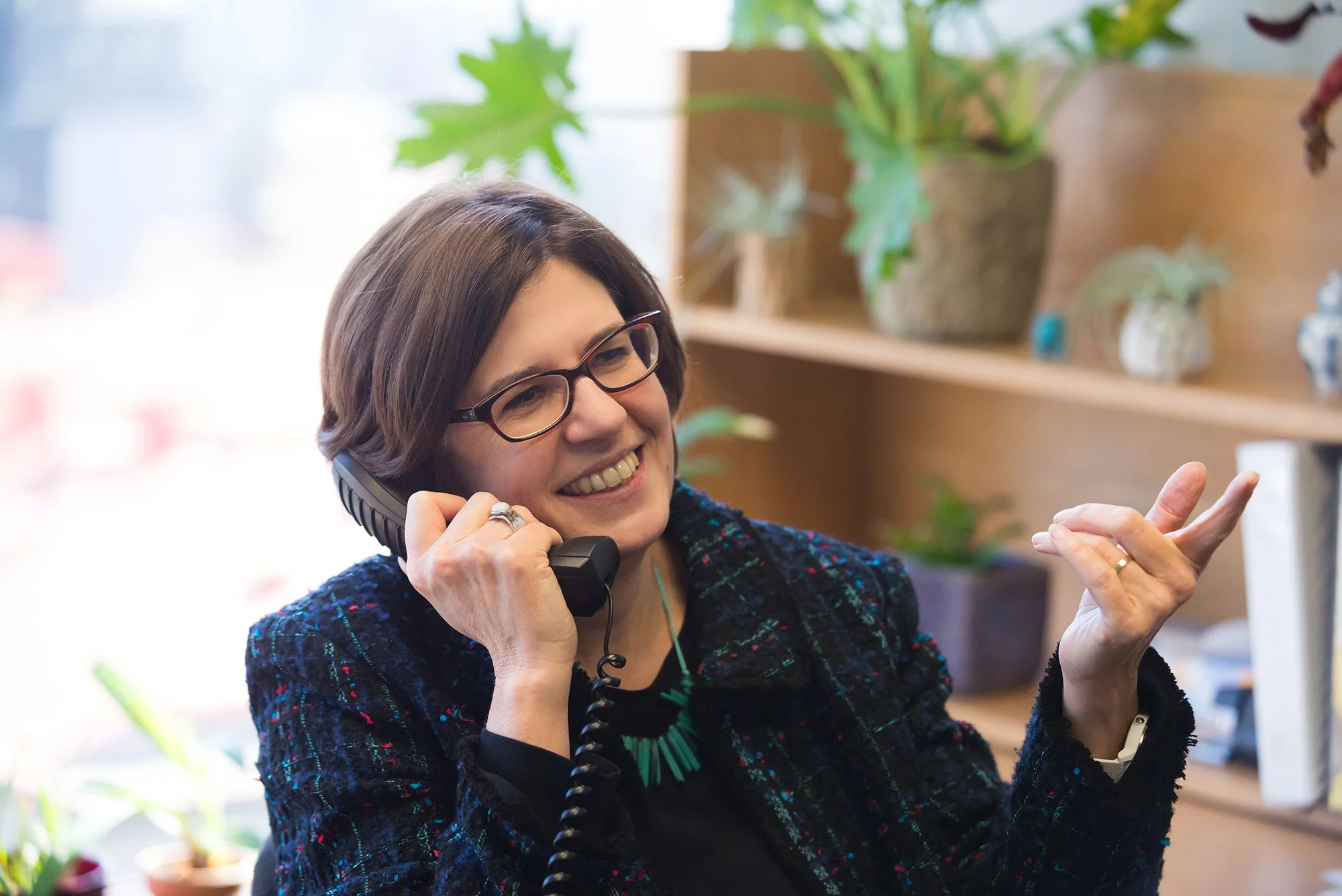 A woman with dark hair, glasses, and a colorful blazer is smiling while talking on a corded landline phone, sitting in a cozy office or home space with plants and decorative objects in the background.