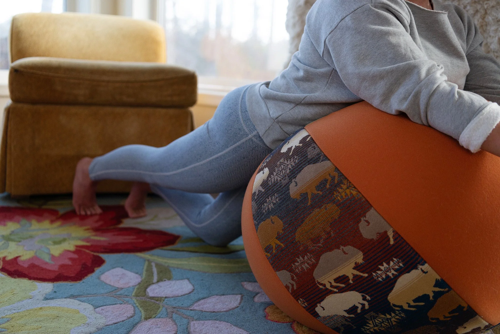 Person pulling a large upholstered footstool with a colorful buffalo plaid pattern, a brown armchair, and a floral area rug in a living room.