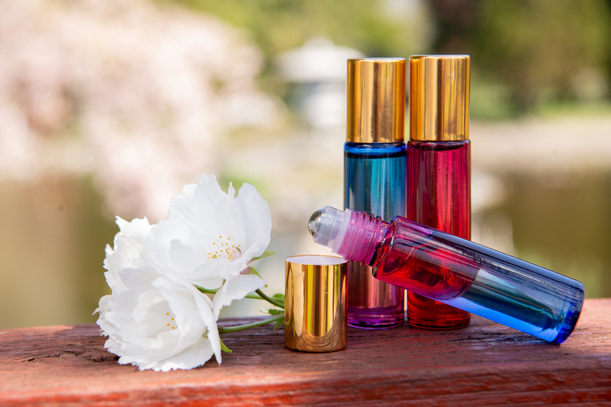 Four colorful perfume bottles and a white flower on a wooden surface outdoors.
