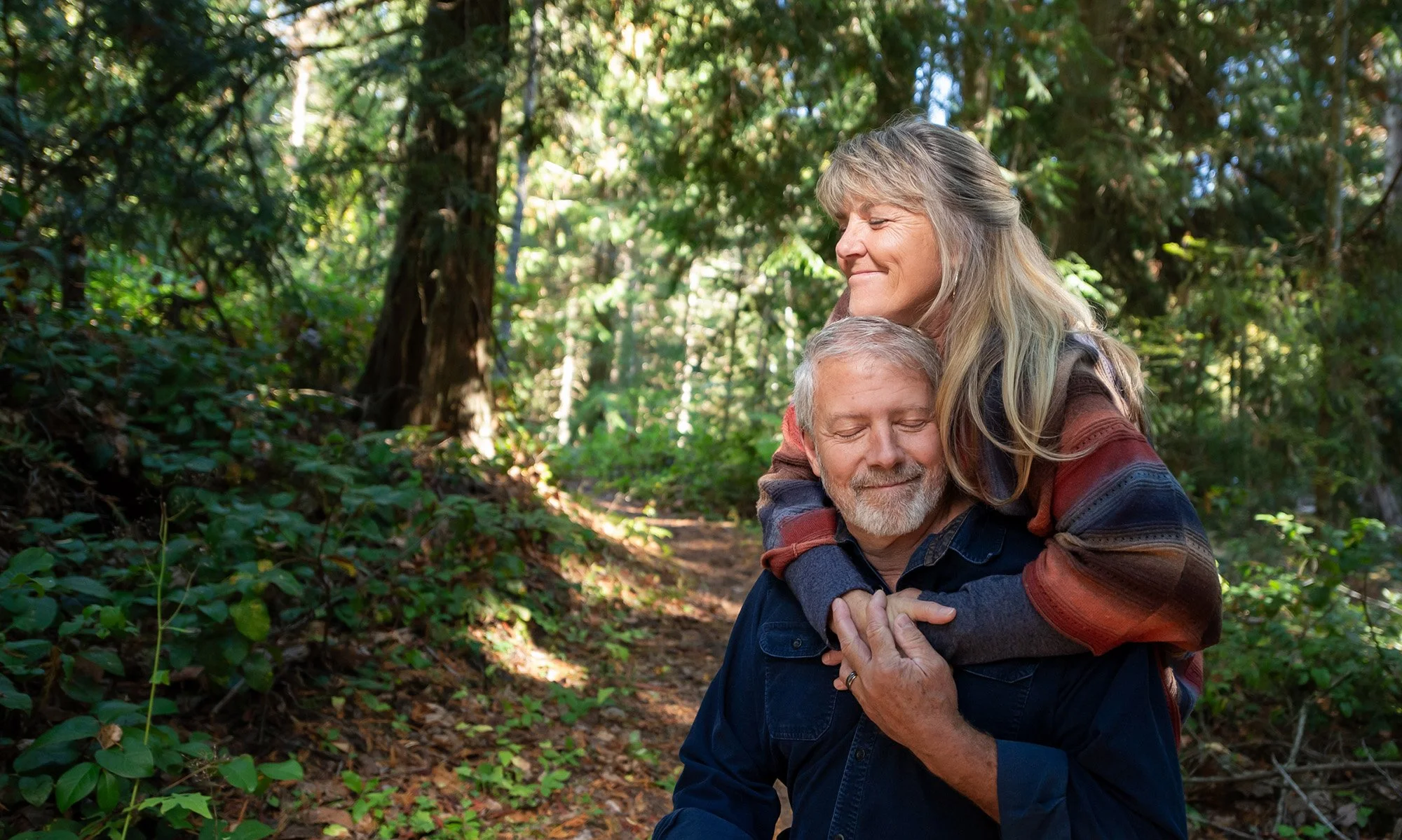 An older man and woman sharing a hug in a forest, both with peaceful and content expressions.